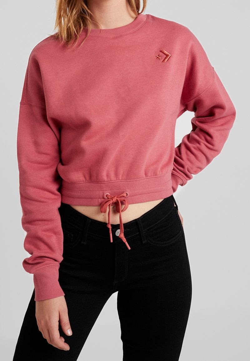 Mujer vestida con un sudadera corta de color rosa polvo con un cordón en la cintura y jeans skinny negros de tiro alto, sobre un fondo liso.