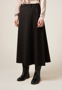 Kvinna klädd i en beige blus, lång svart A-linjeformad kjol och svarta midjekorta stövlar med vit söm, stående mot en enfärgad bakgrund.