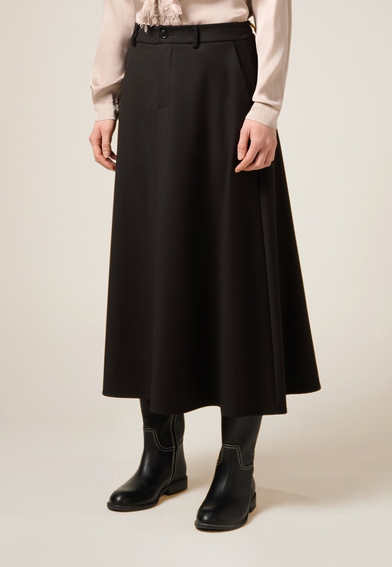 Kvinna klädd i en beige blus, lång svart A-linjeformad kjol och svarta midjekorta stövlar med vit söm, stående mot en enfärgad bakgrund.