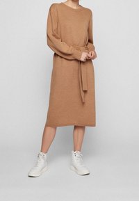 Strickkleid in Camel mit langen Ärmeln, tailliertem Gürtel und gerippten Bündchen. Kombiniert mit weißen, hoch geschnittenen Sneakers.