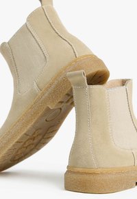 Stivaletti alla caviglia in suede beige con pannelli laterali elasticizzati e linguette di aggancio. Le suole in gomma e i dettagli di cucitura visibili aggiungono texture e struttura.