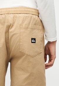 Μπεζ σορτς με ελαστική μέση και πίσω τσέπη που φέρει ένα μικρό ασπρόμαυρο λογότυπο Quiksilver, φορεμένα με λευκό πουκάμισο μακρυμάνικο.