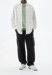 Camisa de cuadros clara abotonada sobre una sudadera verde menta, combinada con pantalones cargo negros. Zapatillas blancas con detalles en azul completan el conjunto.