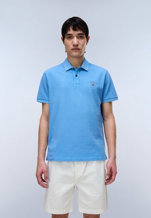 Mann trägt ein hellblaues Polohemd mit kurzen Ärmeln und weiße, knielange Shorts, steht vor einem einfarbigen, hellen Hintergrund.