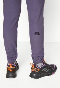 Schwarze Wanderschuhe mit lebhaften orangefarbenen und lila Akzenten. Texturierte Sohlen und reflektierende Streifen. Graue Jogginghosen mit schwarzem Logo-Detail.