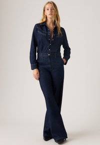 Macacão de jeans escuro com corpete ajustado, mangas longas e pernas largas. Apresenta um colarinho, bolsos no peito e fecho frontal com botões.