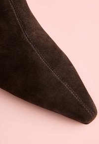 Apdullinātā melna zamša potīšu zābaks ar norādītu purngala formu, gludu tekstūru un redzamām šuvēm, novietots uz maigi rozā fona.