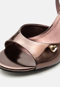 Λαμπερό μπρονζέ σανδάλι με ανοιχτά δάχτυλα, διακοσμημένο με χρυσό καρδιά σε σχήμα διακοσμητικό στοιχείο στο λουράκι και απαλό ροζ εσωτερικό επένδυμα.