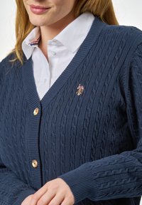 Merino-punottu tummansininen neuletakki, jossa on V-pääntie, kaapelikuvioita, puunappikiinnitys ja pieni brodeerattu logo rinnassa.
