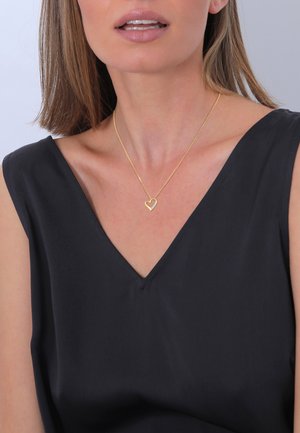 Gul hjerte halskæde med en delikat kæde, båret af en model med en dyb V-udskæring i en sort beklædning. Glat hudtone, subtil makeup og glat hair.