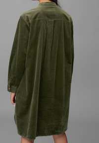 Vista posteriore di una persona che indossa un vestito in velluto a coste verde oliva, largo e a maniche lunghe, con una piega sul retro e maniche ripiegate.