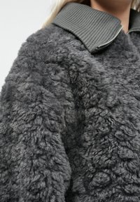 Nærbillede af en grå, tekstureret fleecejakke med ribbet krave og delvis sølvlynlås, båret af en person med blond hår.