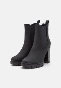 Botas negras de ante hasta el tobillo con un tacón alto y grueso, paneles laterales elásticos y una suela de goma texturizada, que presentan una silueta estilizada.