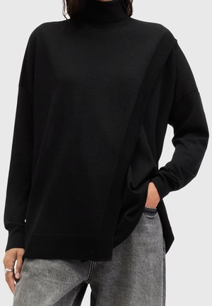 Person, der einen lockeren schwarzen Rollkragenpullover mit subtilen diagonalen Details trägt, kombiniert mit grau gewaschenen Jeans und einer Hand in der Tasche.