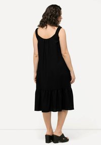 Schwarzes ärmelloses Kleid mit einem Rüschensaum und gerafftem Rücken, aus leichtem Stoff gefertigt. Getragen mit strukturierten schwarzen Wedge-Sandalen.