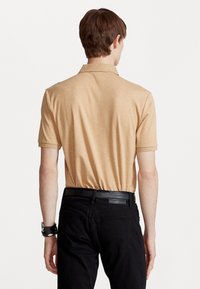 Polo shirt corto de manga corta de color beige con una textura suave y un collar clásico, combinado con jeans negros y un cinturón negro. Vista desde la espalda.