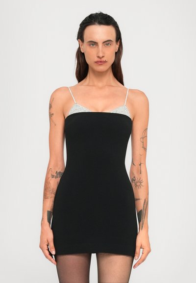 Mujer con cabello largo y oscuro que lleva un vestido mini sin mangas negro sobre una blusa de tirantes plateados, exhibiendo múltiples tatuajes en ambos brazos.
