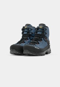Des bottes de randonnée bleues en suede et en matériaux synthétiques, dotées de semelles en caoutchouc noir, de capuchons de protection renforcés et de lacets avec des accents rouges.