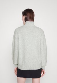 Za duży, jasnoszary golf z miękkiego dzianego materiału, z prążkowanymi mankietami i dołem. Noszony z ciemną spódnicą, widok od tyłu.