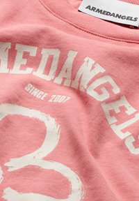 Roze katoenen t-shirt met witte tekst die "ARMEDANGELS" en "SINCE 2007" zegt, met een groot wit circulair grafisch ontwerp.