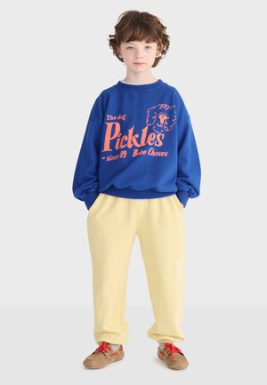 Bambino con capelli ricci castani che indossa una felpa blu con scritta arancione e pantaloni della tuta gialli, in piedi con le mani nelle tasche.