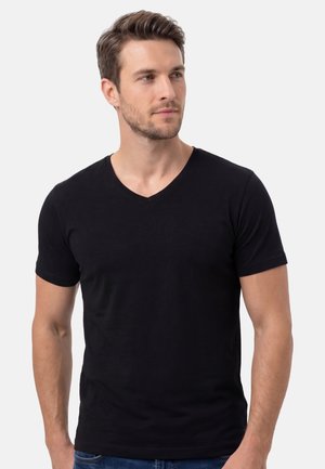 Homme aux cheveux bruns courts portant un T-shirt noir à col en V, debout devant un fond clair uni, regardant sur le côté.