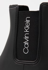 Bottines Chelsea noires en matériau caoutchouteux lisse, avec un panneau latéral élastique et un logo "Calvin Klein" blanc proéminent sur un tissu noir texturé.