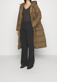 Mujer vistiendo un abrigo largo acolchado marrón sobre una blusa negra de cuello en V, pantalones anchos negros y zapatos negros, de pie contra un fondo blanco liso.
