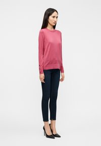 Εφαρμοστό ροζ πουλόβερ με μακριά μανίκια, στρογγυλή λαιμόκοψη και διακοσμητικά κουμπιά στις μανσέτες, συνδυασμένο με στενά σκούρα τζιν και μαύρες ψηλοτάκουνες γόβες.