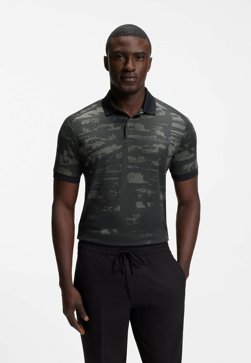 Homme portant un polo noir à manches courtes avec un motif numérique abstrait gris et un pantalon noir à cordon, debout sur fond blanc.
