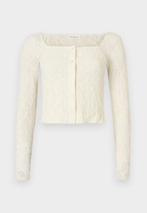 Cardigan court en dentelle crème à manches longues, avec col carré et quatre boutons sur le devant, présenté sur un fond blanc uni.