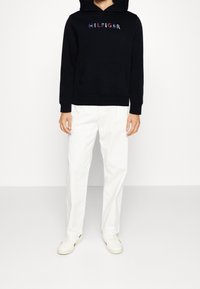Felpa nera con logo "HILFIGER" in rosso, bianco e blu, dotata di tasca frontale. Abbinata a pantaloni bianchi a gamba dritta e sneakers bianche.