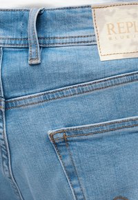 Ανοιχτό μπλε τζιν παντελόνι με εμφανή πίσω τσέπη, θηλιά για ζώνη και λευκή ορθογώνια ετικέτα μάρκας ραμμένη κοντά στη μέση.