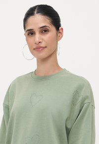 Zeleno pulover iz mehkega blaga, okrašen s kamenčki v obliki srca na sprednji strani in s krožnim izrezom.