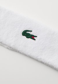 Weißes Handtuchstoff-Haarband mit weicher Textur. Verfügt über ein grünes Krokodil-Logo mit roten Akzenten. Entwickelt für Komfort und sportliche Nutzung.
