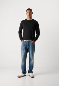Mann steht mit Händen in den Taschen, trägt einen schwarzen Pullover über einem weißen Shirt, blaue Jeans und weiße Sneakers vor weißem Hintergrund.