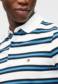 Κοντινό πλάνο ενός ατόμου που φοράει λευκό πουκάμισο πόλο με οριζόντιες μπλε και μαύρες ρίγες και ένα μικρό ορθογώνιο λογότυπο στο στήθος.