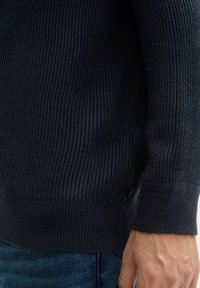 Pull en maille côtelée bleu foncé, présentant une surface texturée, un col rond et des fentes sur les côtés au niveau de l'ourlet pour un ajustement décontracté.