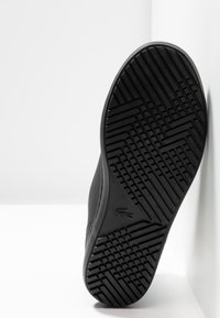Semelle de chaussure noire avec motif géométrique texturé, inclinée sur fond blanc, montrant un design détaillé de l’adhérence et un petit logo.