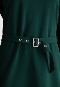 Zelené šaty s hladkou textúrou, s opaskom s prievlakmi a kovovou sponou, zdôrazňujúcim štruktúrovaný dizajn.