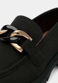 Chaussure mocassin noire avec semelle épaisse, ornée d’un détail décoratif en chaîne entrelacée dorée et noire sur le dessus.