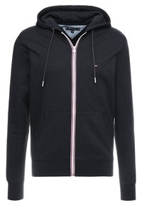 Felpa nera con zip realizzata in tessuto morbido, dotata di tasca frontale, cappuccio con coulisse e dettagli a strisce rosse e bianche lungo la zip.