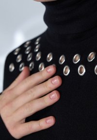 Schwarzer Rollkragenpullover mit metallischen Ösenakzenten am Kragen, versehen mit Rippstruktur und taillierter Passform. Eine Hand ruht auf dem Stoff.