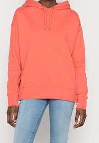 Modello che indossa un felpa corallo con una vestibilità comoda, abbinata a jeans azzurri chiari. L'attenzione è rivolta all'abbigliamento e alla postura del modello.