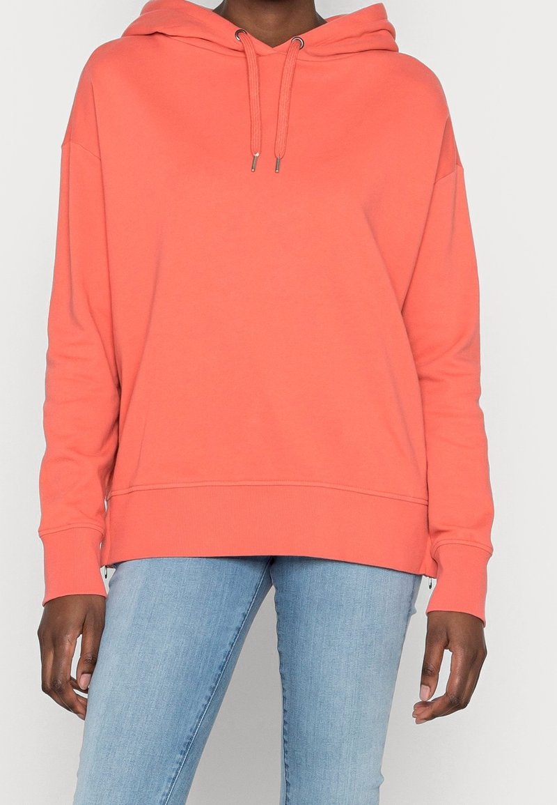 Modello che indossa un felpa corallo con una vestibilità comoda, abbinata a jeans azzurri chiari. L'attenzione è rivolta all'abbigliamento e alla postura del modello.