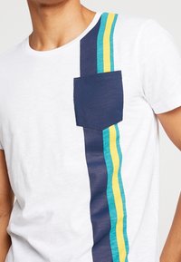 Camiseta blanca con un bolsillo en el pecho en azul marino y rayas verticales en verde agua, amarillo y azul oscuro en el lado izquierdo. Material de algodón, corte informal.