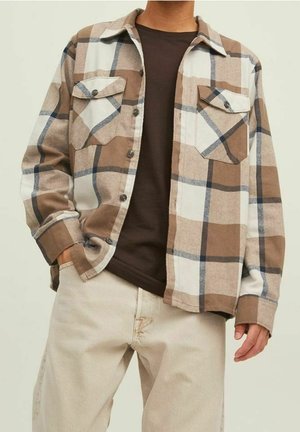 Mann trägt ein braun-cremefarbenes kariertes Hemd über einem dunkelbraunen T-Shirt mit hellbeigen Hosen, Hand in der Tasche.