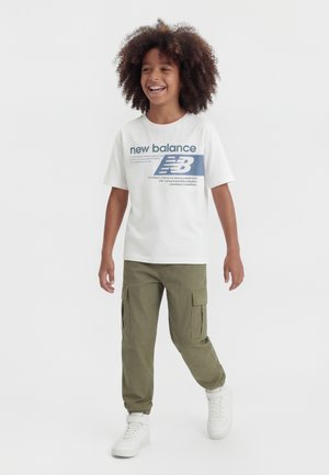 Niño sonriente con cabello rizado camina hacia adelante vistiendo una camiseta blanca de New Balance, pantalones cargo oliva y zapatillas blancas sobre un fondo liso.