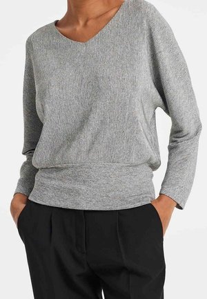 Modèle portant un haut gris côtelé à manches longues avec un large décolleté et un pantalon noir, mains dans les poches.