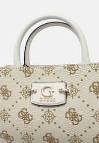 Sac à main en textile blanc avec un motif doré, présentant une forme structurée, des poignées blanches doubles et un fermoir en métal argenté avec les marques "G" et "GUESS".